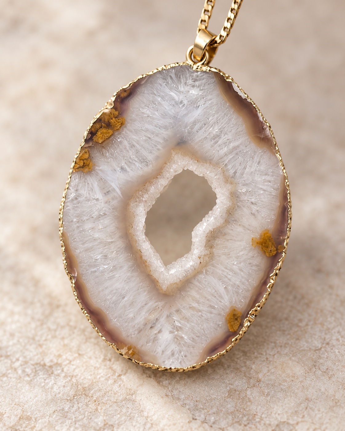 A gold bezel pendant necklace featuring a polished agate slice with a white and brown crystalline center and small gold inclusions, shown on a textured beige surface.