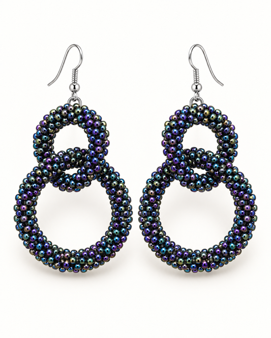 A pair of large, double-loop dangle earrings made from iridescent seed beads that shift between black, purple, and blue, suspended from silver fishhook wires on a plain white background.
