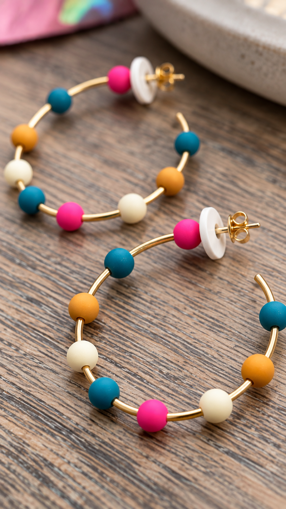 Gold hoop earrings with colorful spacer beads in pink, teal, orange, and cream, displayed on a dark wood surface.