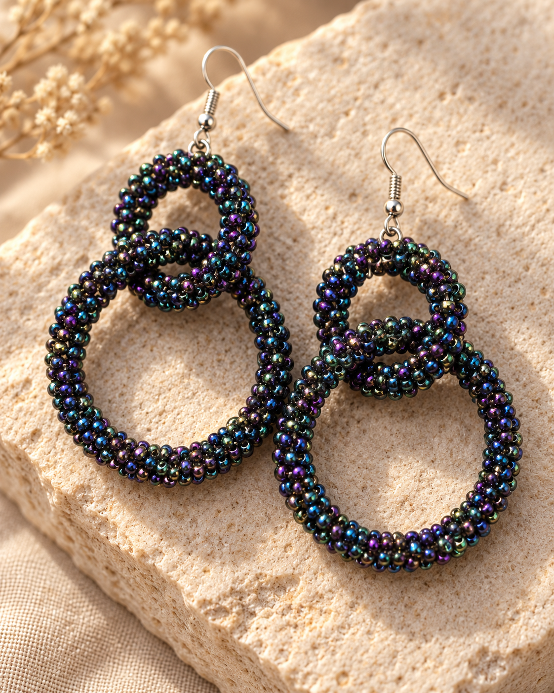 A pair of large beaded hoop earrings with silver fishhook wires, displayed on a textured beige stone surface with dried flowers in the background.