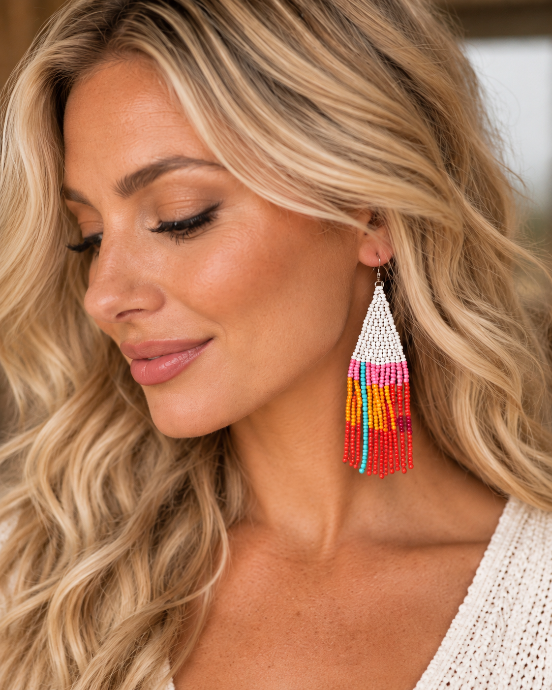 A close-up of a woman wearing a long, beaded tassel earring. The earring features a white beaded triangular top with colorful dangling tassels in pink, orange, yellow, and blue.