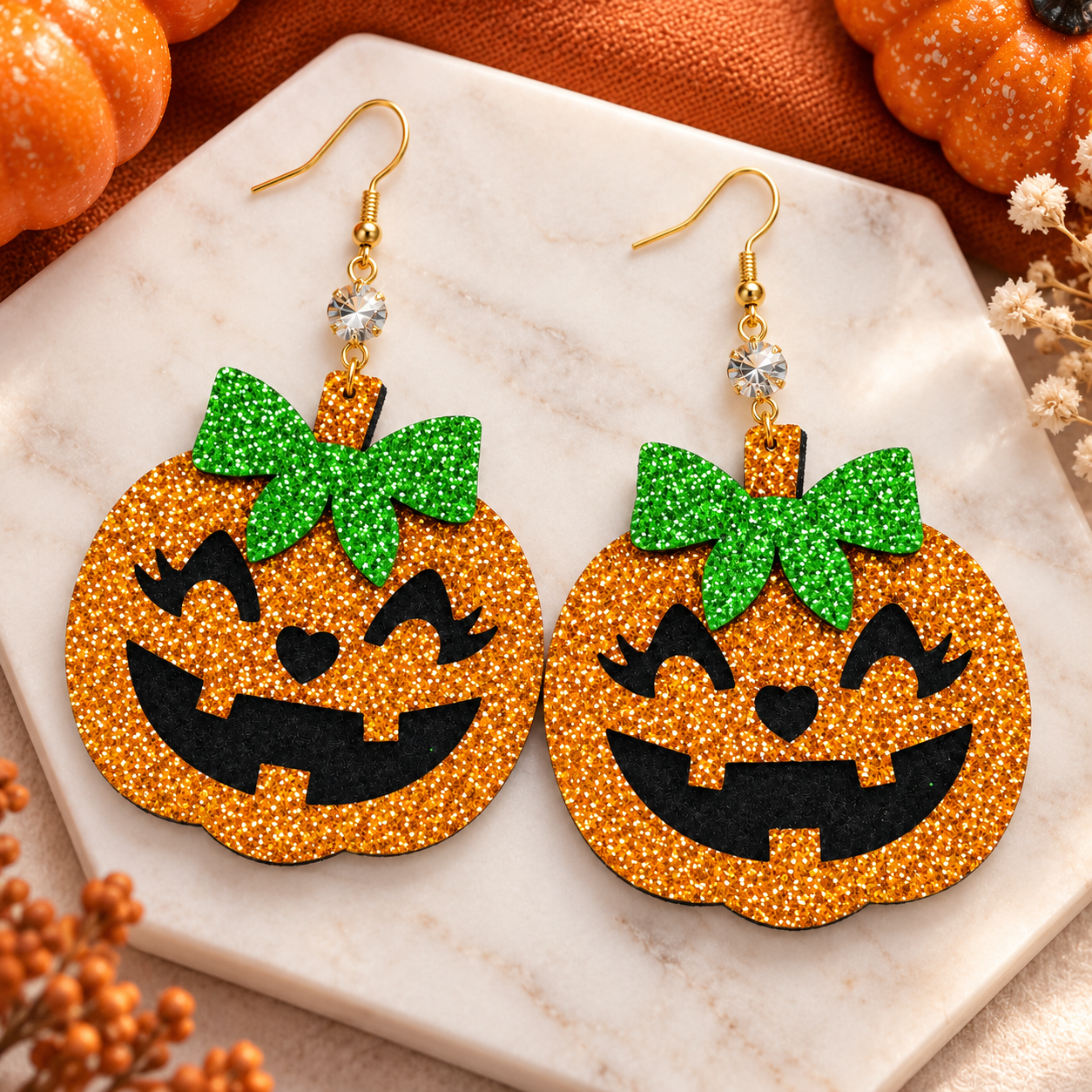 A pair of Halloween pumpkin-shaped dangle earrings with glittery orange bodies, green glitter bows, black jack-o'-lantern faces, and gold fishhook wires. They are displayed on a white marble tray with autumn props including orange fabric, small pumpkins, and dried flowers.