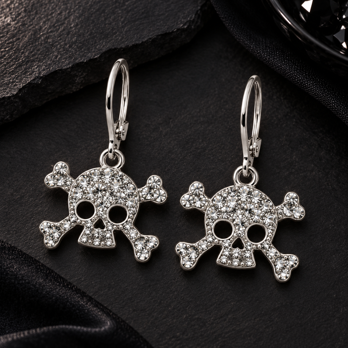 A pair of silver-toned skull and crossbones drop earrings paved with clear rhinestones, displayed on a dark textured surface.