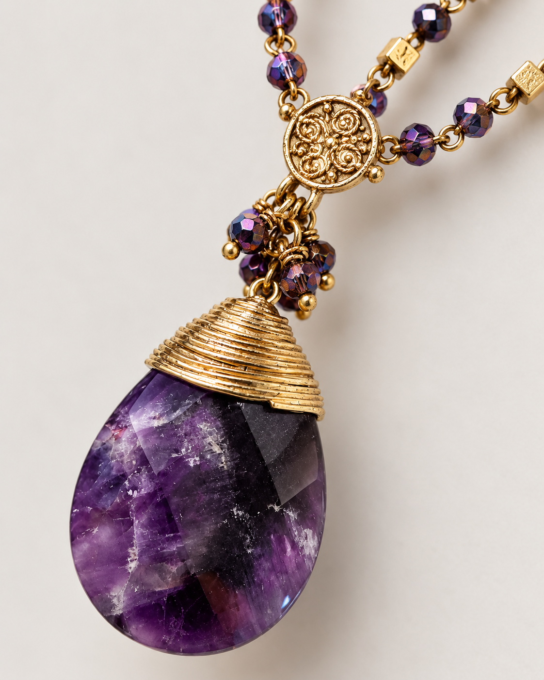 A close-up of a teardrop-shaped purple gemstone pendant with textured gold metal wrapping, hanging from a gold chain with faceted purple beads on a light gray background.