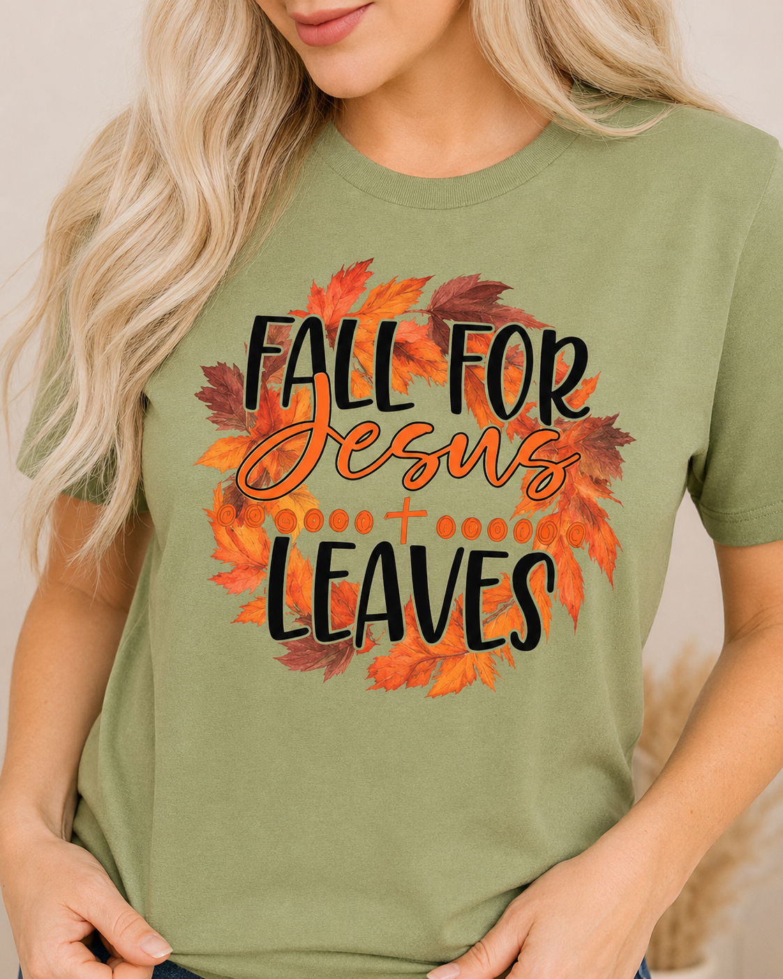 A woman wearing an olive green t-shirt with a graphic that reads 'FALL FOR Jesus LEAVES' in black and orange script, surrounded by a circular wreath made of orange and brown autumn leaves.