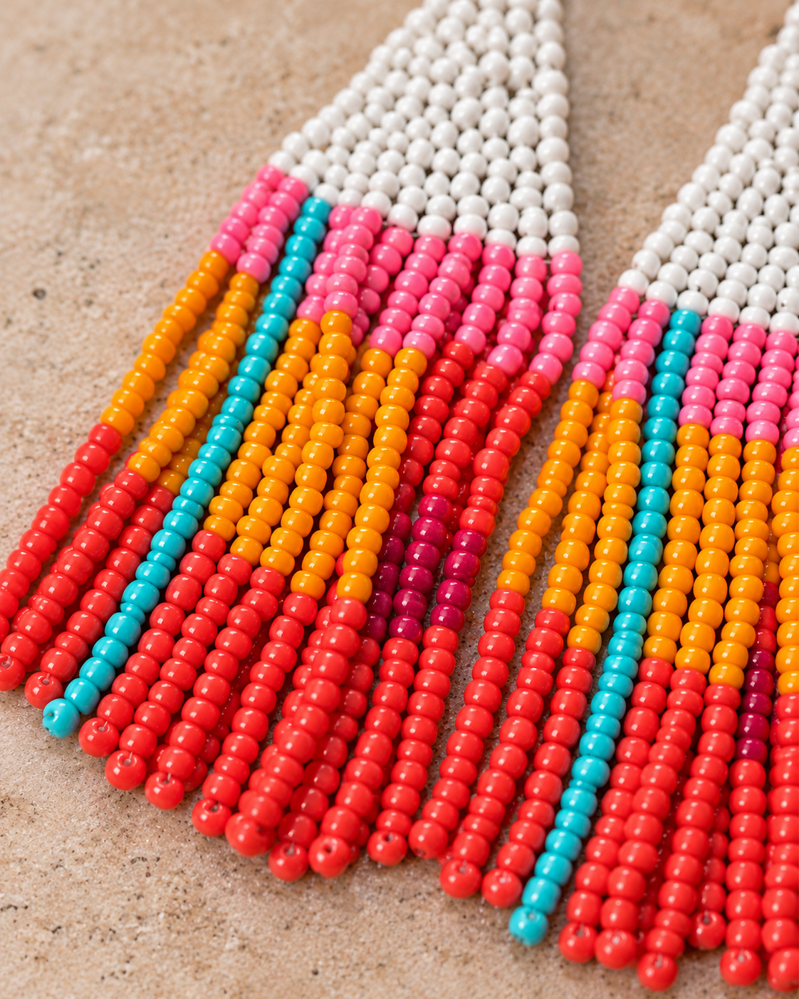 Close-up of a pair of beaded fringe earrings laid flat on a textured beige surface. The earrings have white bead tops, then vertical rows of pink, orange, red, and turquoise beads forming a striped fringe pattern.
