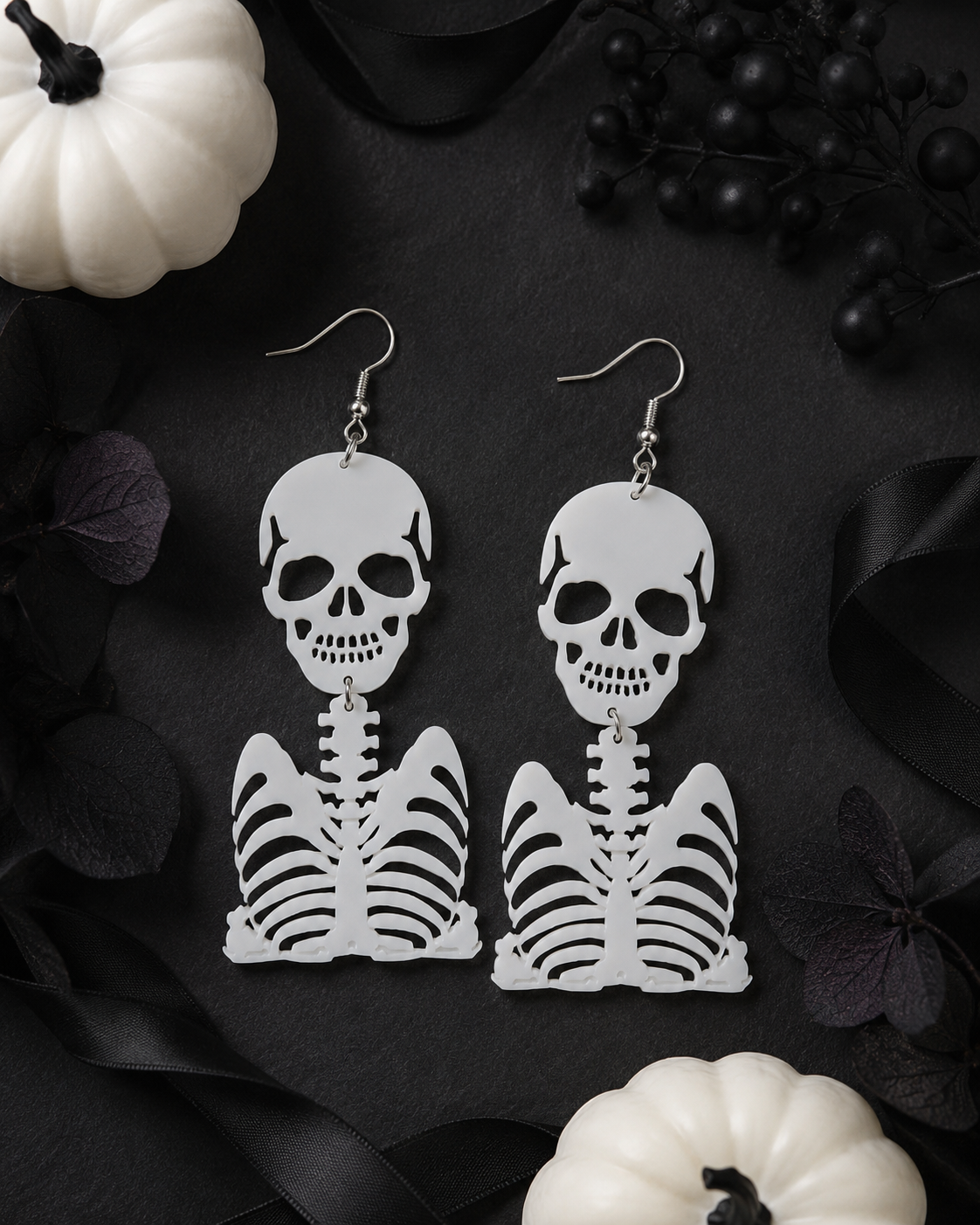 White skeleton earrings styled on a black background with ribbon, dark florals and white pumpkins