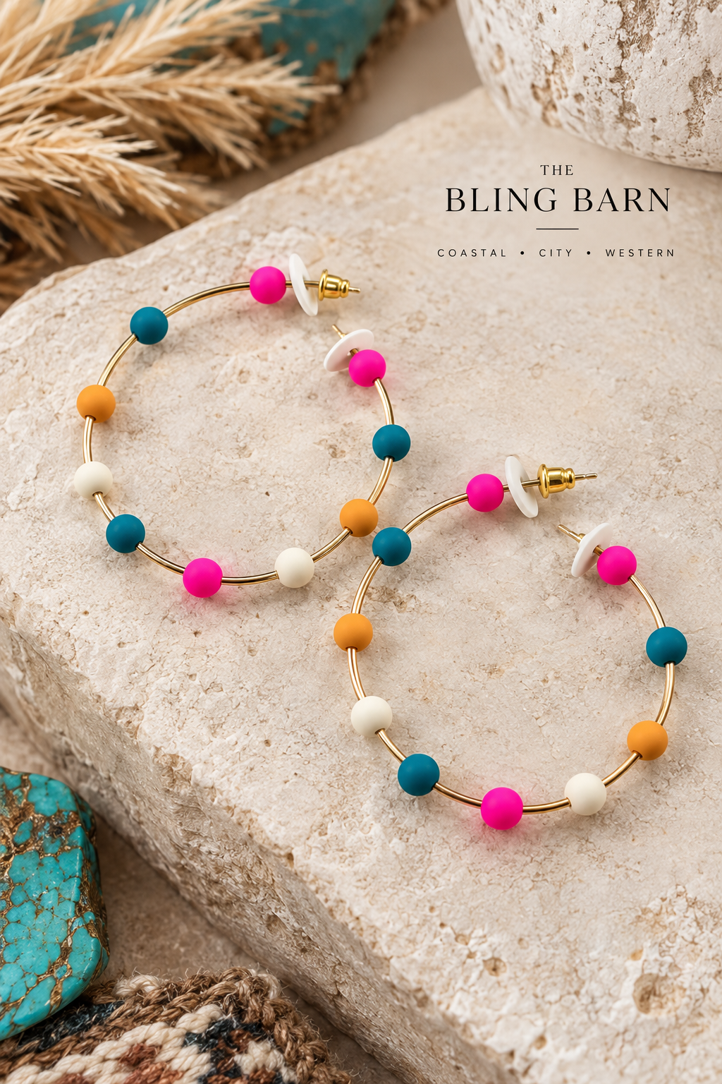 A pair of gold hoop earrings with colorful beaded accents in pink, teal, orange, and white, displayed on a textured stone surface with boho-style props. The brand name 'The Bling Barn' is visible in the upper right.
