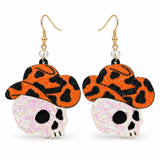 A pair of dangling earrings featuring glittery orange and black patterned cowboy hats with white glitter skulls underneath, suspended from gold hooks on a white background.