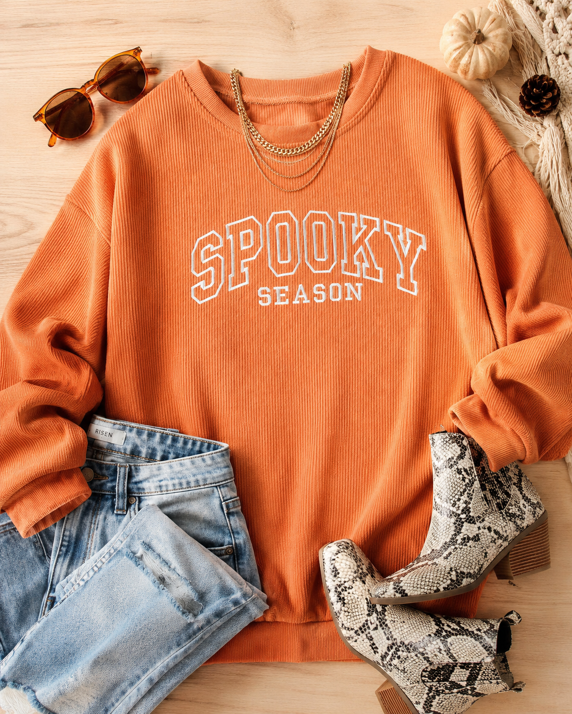 An orange ribbed crewneck sweatshirt with 'SPOOKY SEASON' printed in white varsity letters, styled in a flat lay with light-wash jeans, snakeskin ankle boots, sunglasses, a gold chain necklace, and a knit scarf on a light wood background.