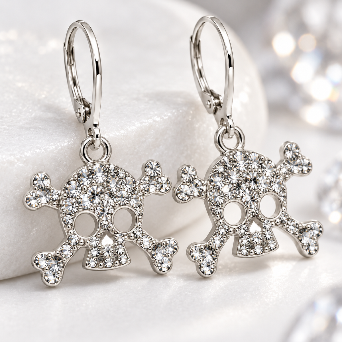 A pair of silver-toned skull and crossbones drop earrings paved with clear rhinestones, resting on a white textured surface with a soft, blurred background of sparkling lights.