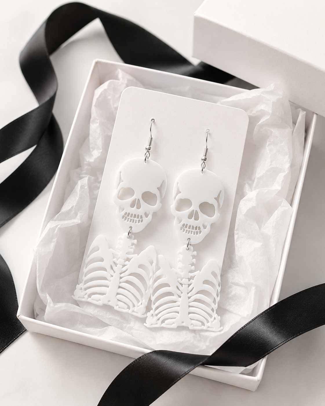 White skull and rib cage earrings displayed on a jewelry card in a gift box with black ribbon