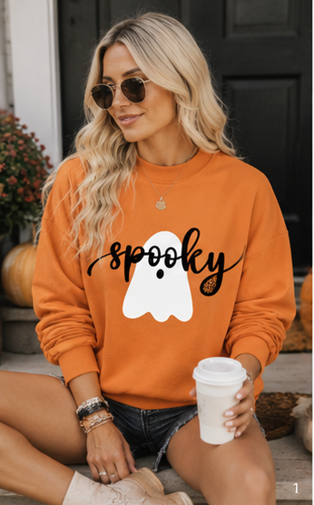 A woman wearing an orange crewneck sweatshirt with a 'spooky' graphic featuring a white ghost and a small acorn, sitting outdoors and holding a white coffee cup.