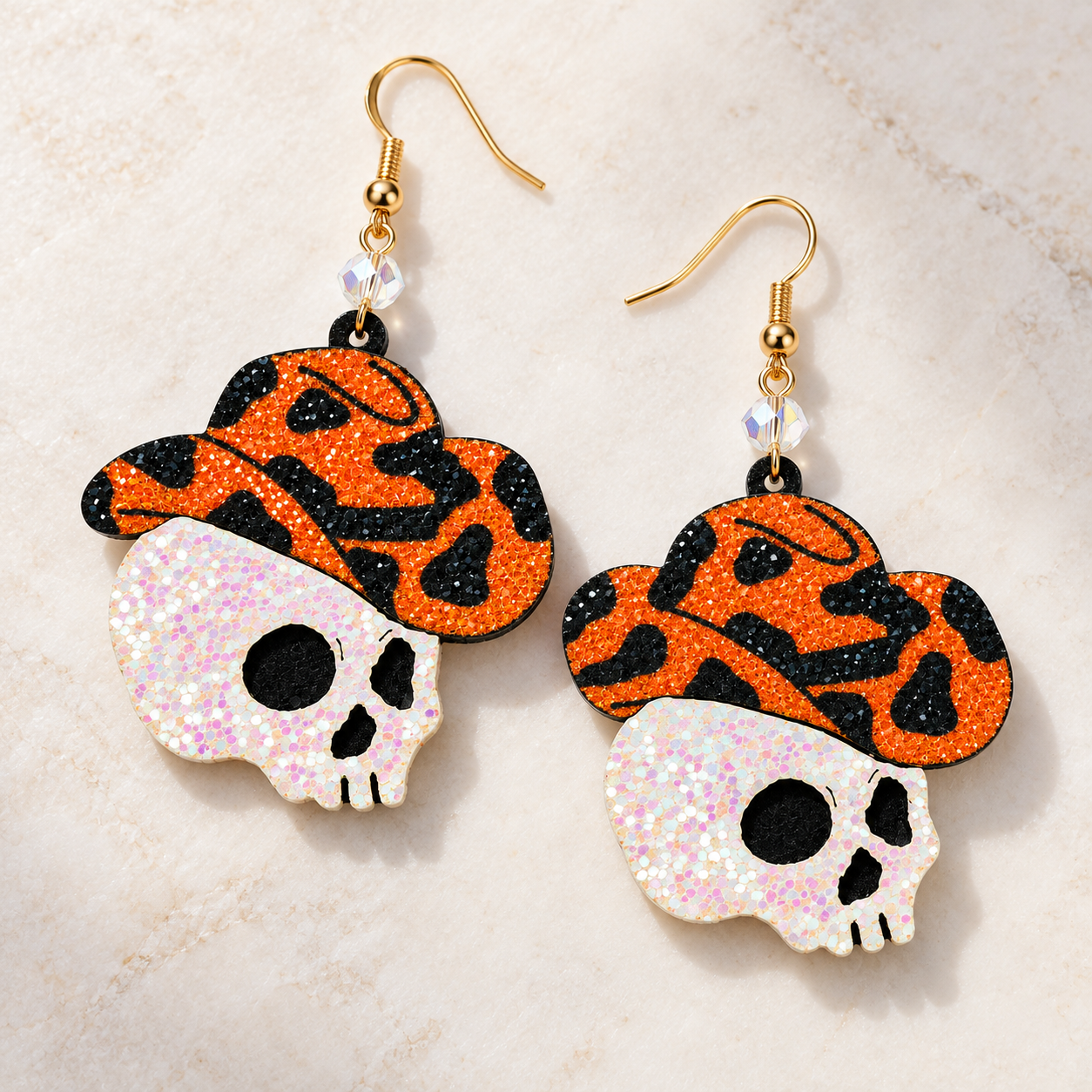 A pair of dangling earrings with glittery orange and black leopard-print hats and white glitter skulls, displayed on a light beige marble background.