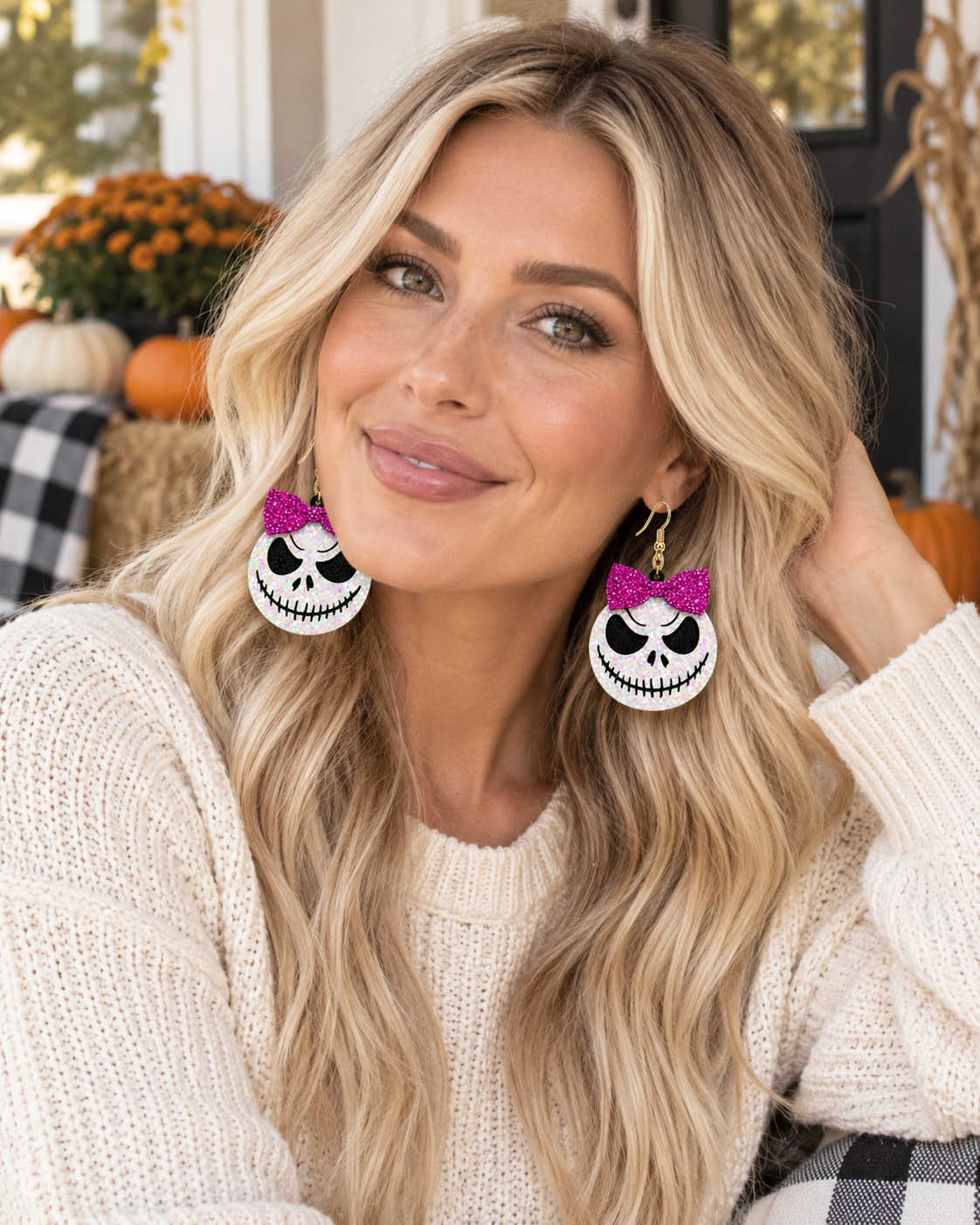 A woman with long blonde hair wearing large white and black skull-shaped drop earrings with bright pink bows, posed in a cozy fall-themed setting with pumpkins and autumn decor in the background.