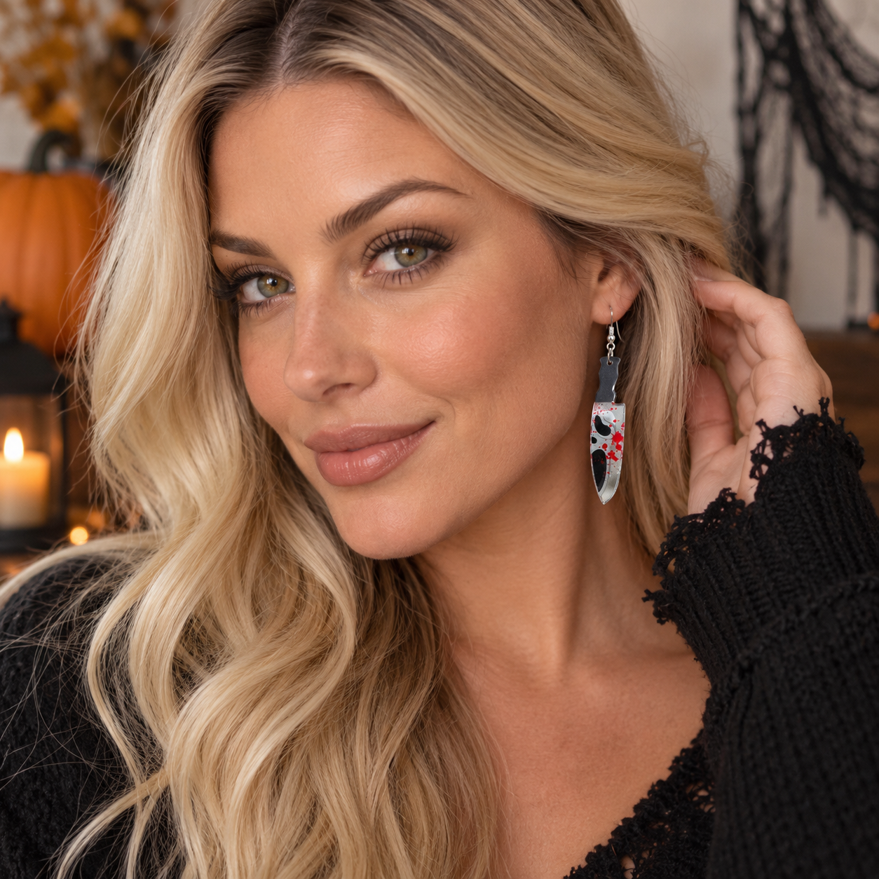 A close-up lifestyle shot of a woman with blonde hair wearing a long silver dangle earring decorated with a black cat illustration and red floral accents, set against a warm, Halloween-themed background.