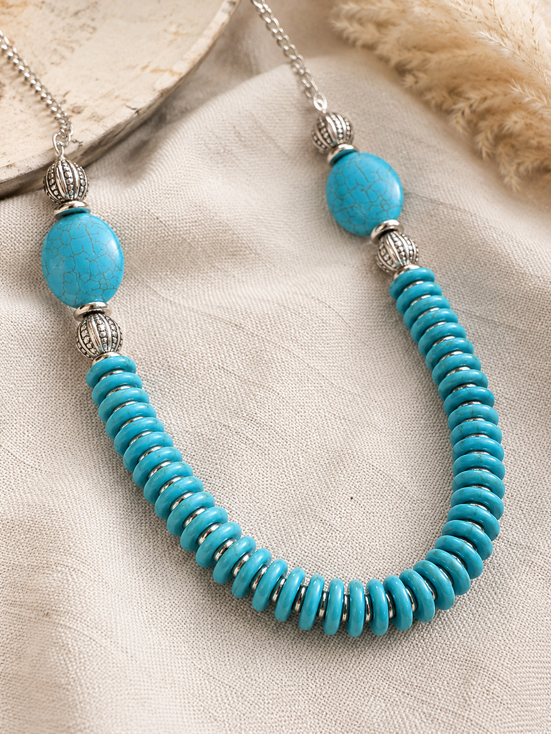 A turquoise and silver beaded necklace displayed on a textured beige fabric background.