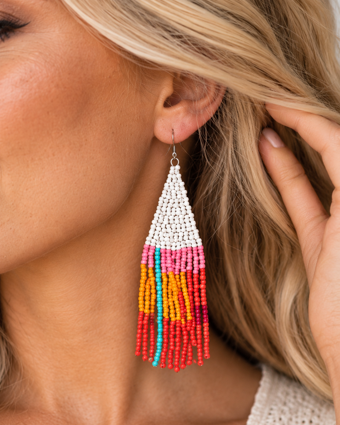A close-up of a woman wearing a long beaded tassel earring. The earring features a white beaded triangular top with colorful fringe strands in pink, orange, yellow, and turquoise red.