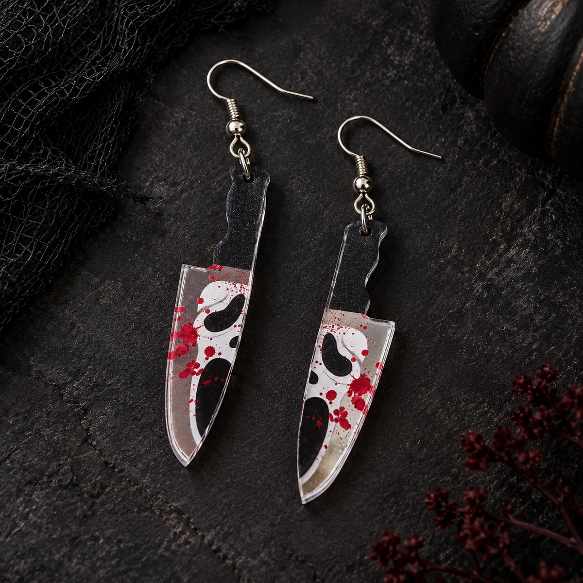 A pair of dangling knife-shaped earrings with a horror mask design and red blood splatter, displayed on a dark textured surface.