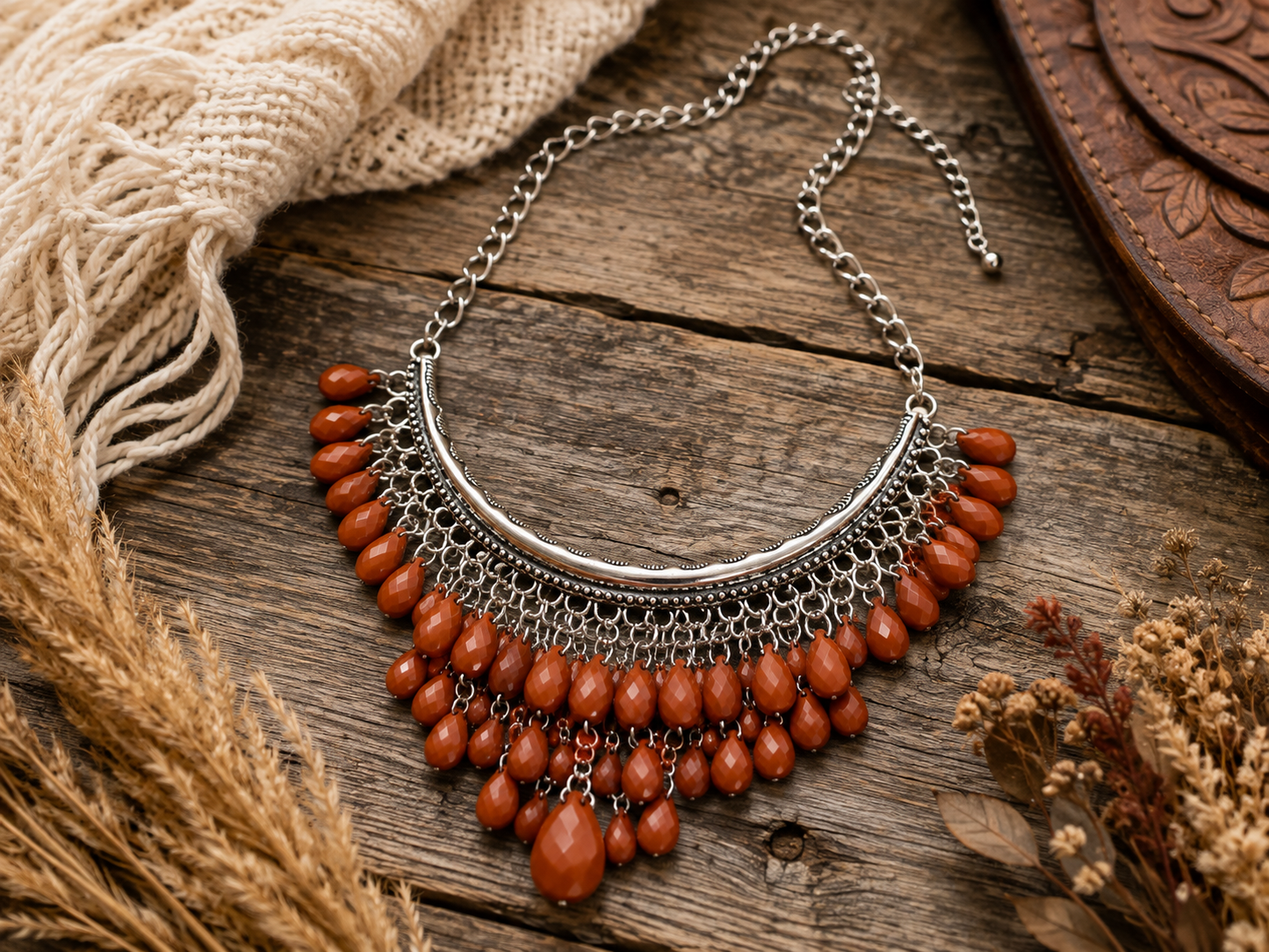 A bohemian-style statement necklace with a silver-toned crescent chain and multiple tiers of faceted terracotta beads, displayed on a rustic wooden surface with dried grass and a woven textile.