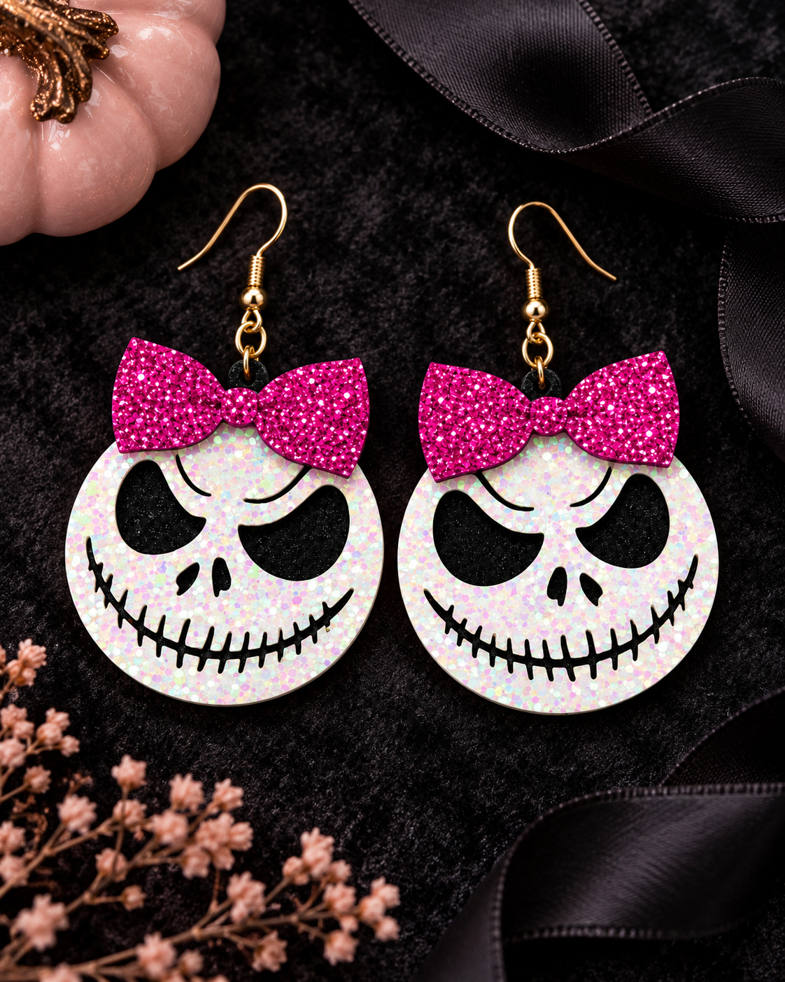 A pair of gothic-style dangle earrings featuring Jack Skellington's face in white glitter, complete with a bright pink glitter bow and gold fishhook ear wires, displayed on a black velvet background with decorative ribbons and dried flowers.