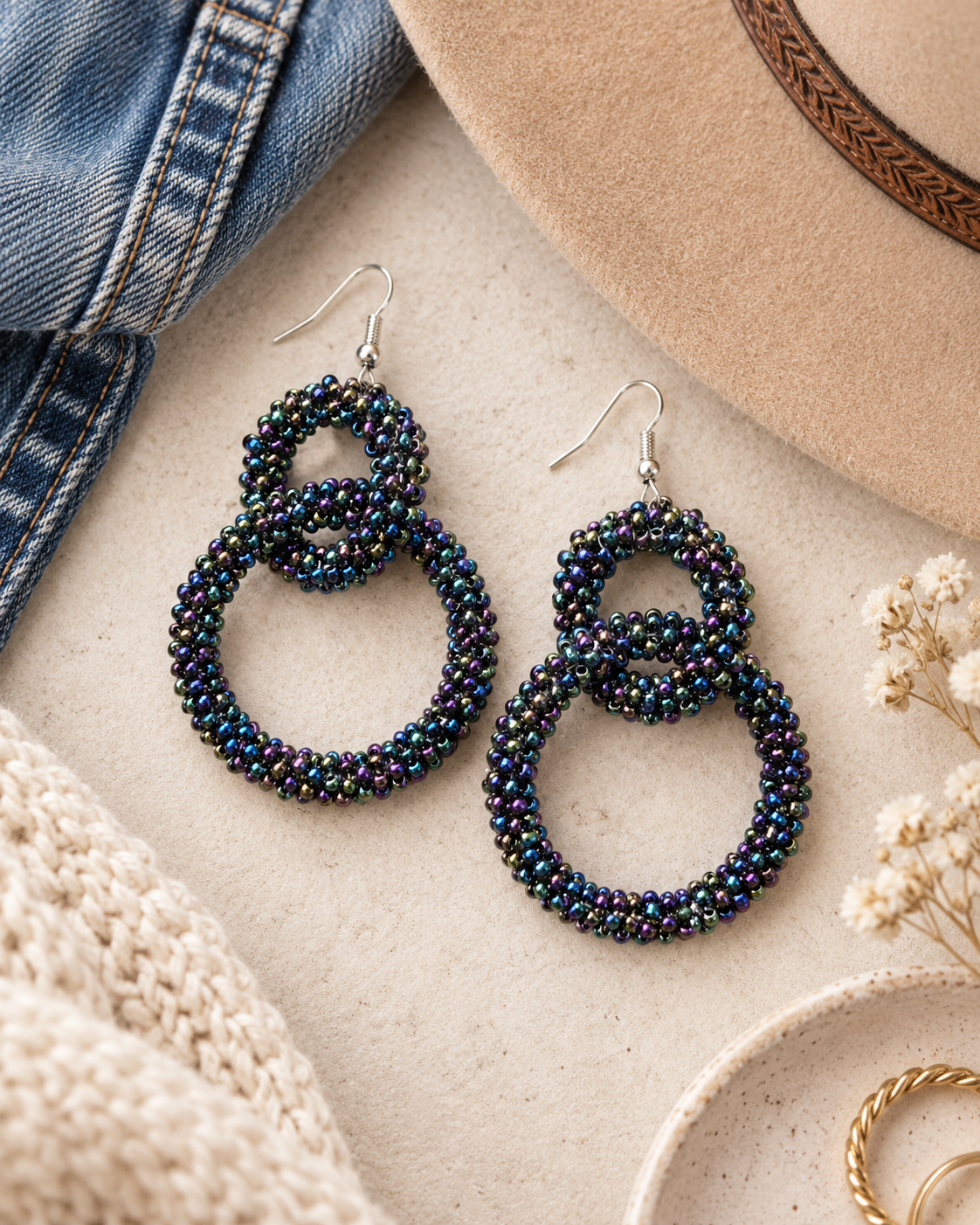 A pair of beaded dangle earrings with interlocking double-ring designs, featuring iridescent beads that shift between dark blue, green, and purple. The earrings are displayed in a styled flat-lay arrangement on a textured beige surface, surrounded by a denim jacket, a tan hat, a knit sweater, and dried flowers.