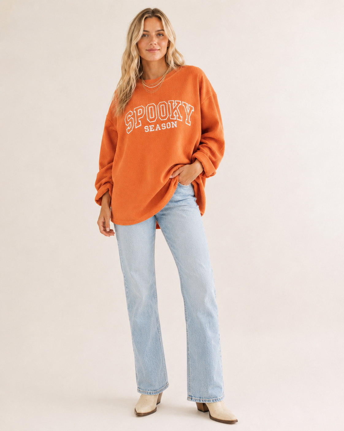 A woman modeling an orange crewneck sweatshirt with 'SPOOKY SEASON' printed in white on the front, paired with light-wash blue jeans and beige ankle boots, against a plain beige studio background.