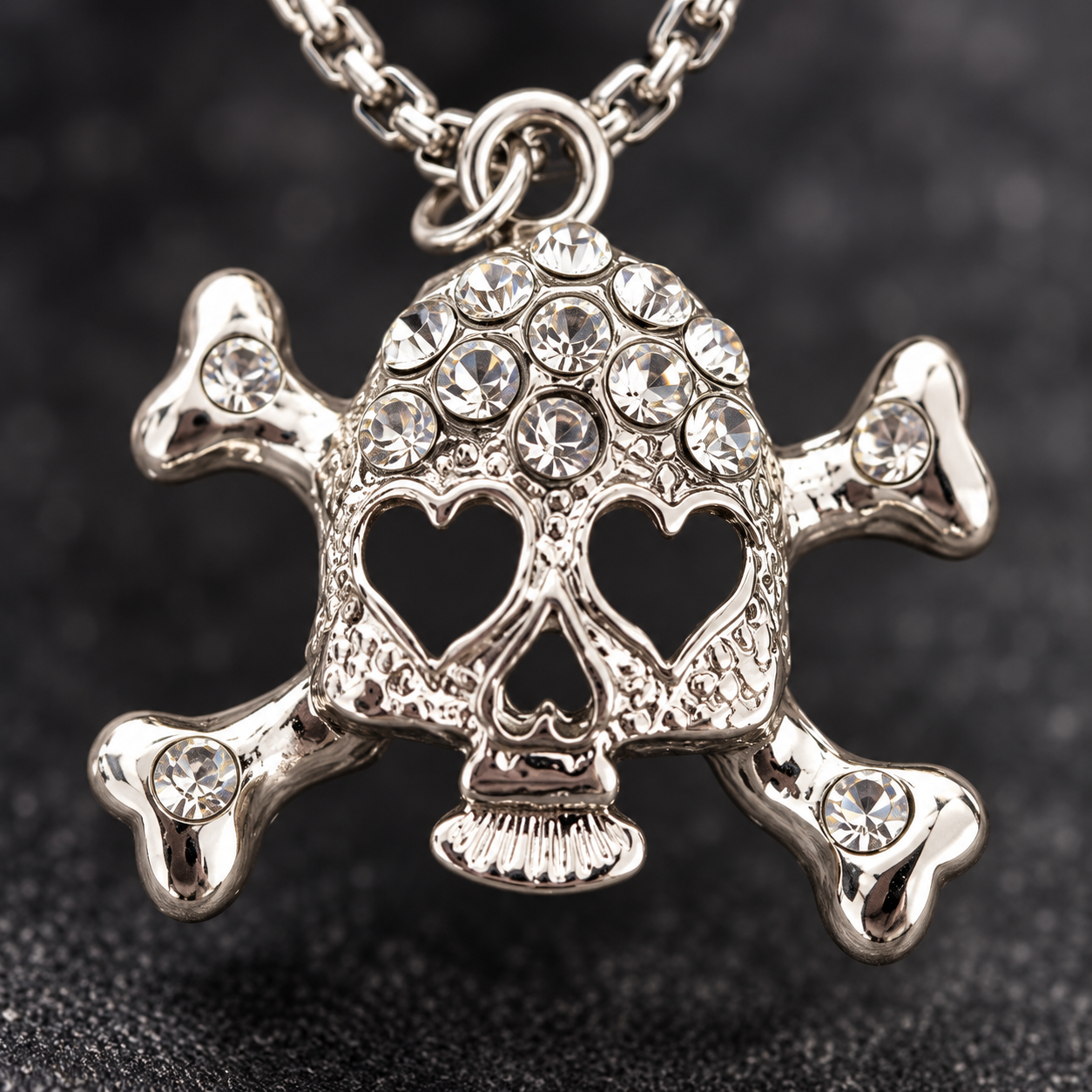 A silver-toned skull and crossbones pendant necklace adorned with clear rhinestones, shown close-up against a dark textured background.