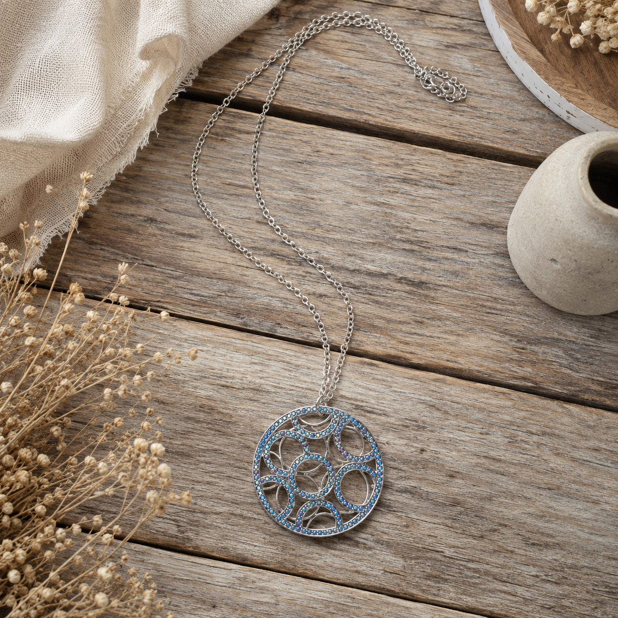 A silver-toned circular pendant necklace with intricate cutout detailing and light blue stones, displayed on a rustic wooden surface with dried flowers and a small ceramic vase.