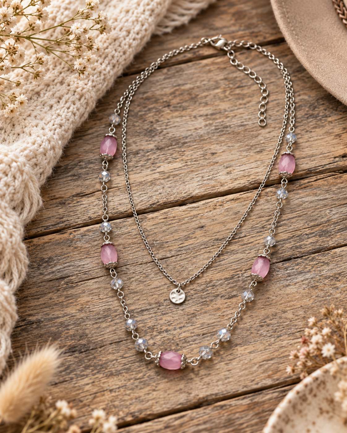 A double-strand silver-toned necklace with pink cabochon beads and clear faceted beads, styled on a rustic wooden surface with a cream knit blanket and dried flowers.