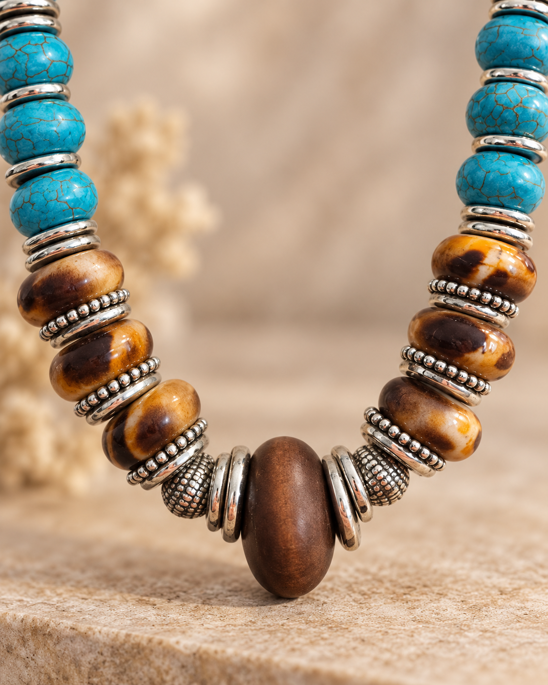 A close-up of a bohemian beaded necklace featuring turquoise, brown tiger's eye, and polished wood beads separated by textured silver spacer rings, displayed on a neutral beige surface.