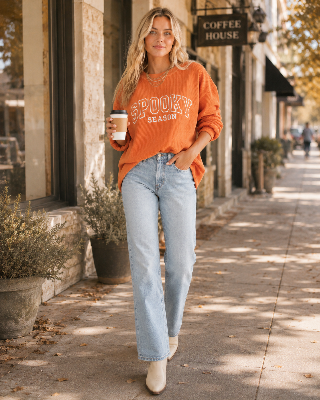 A woman walking on a city sidewalk wearing an orange crewneck sweatshirt with 'SPOOKY SEASON' printed in white across the chest, paired with light blue jeans and white ankle boots, holding a coffee cup.