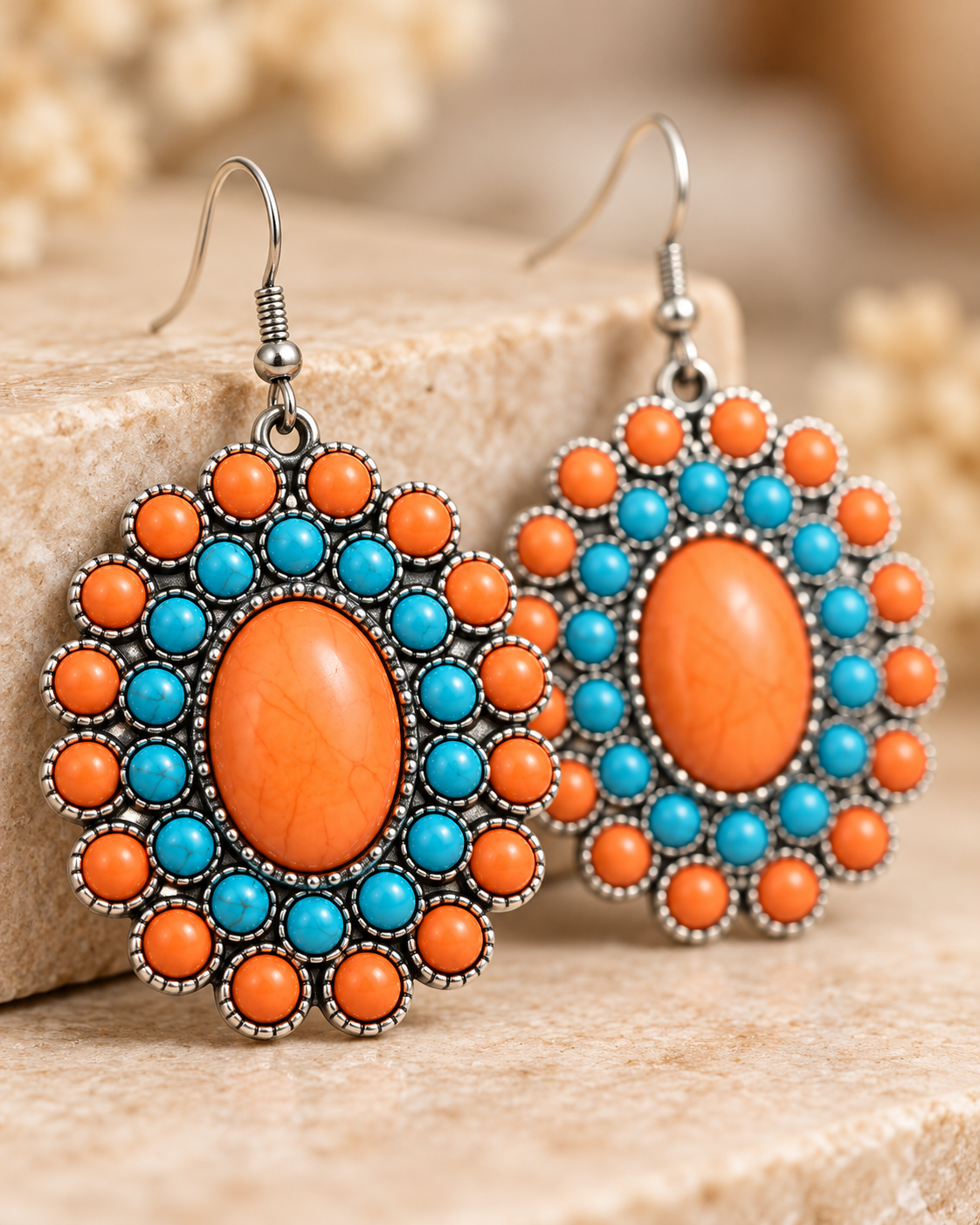A pair of bohemian-style dangle earrings with oval orange centers, surrounded by rings of turquoise blue and orange round beads, set in silver-tone metal, resting on a textured beige stone surface.