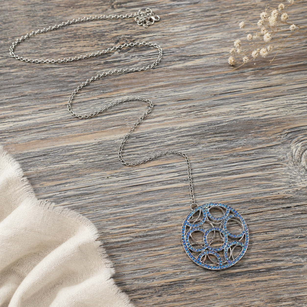 A silver-toned chain necklace with a circular pendant featuring blue floral cutout details, displayed on a rustic wood surface with a cream fabric corner and dried flowers.