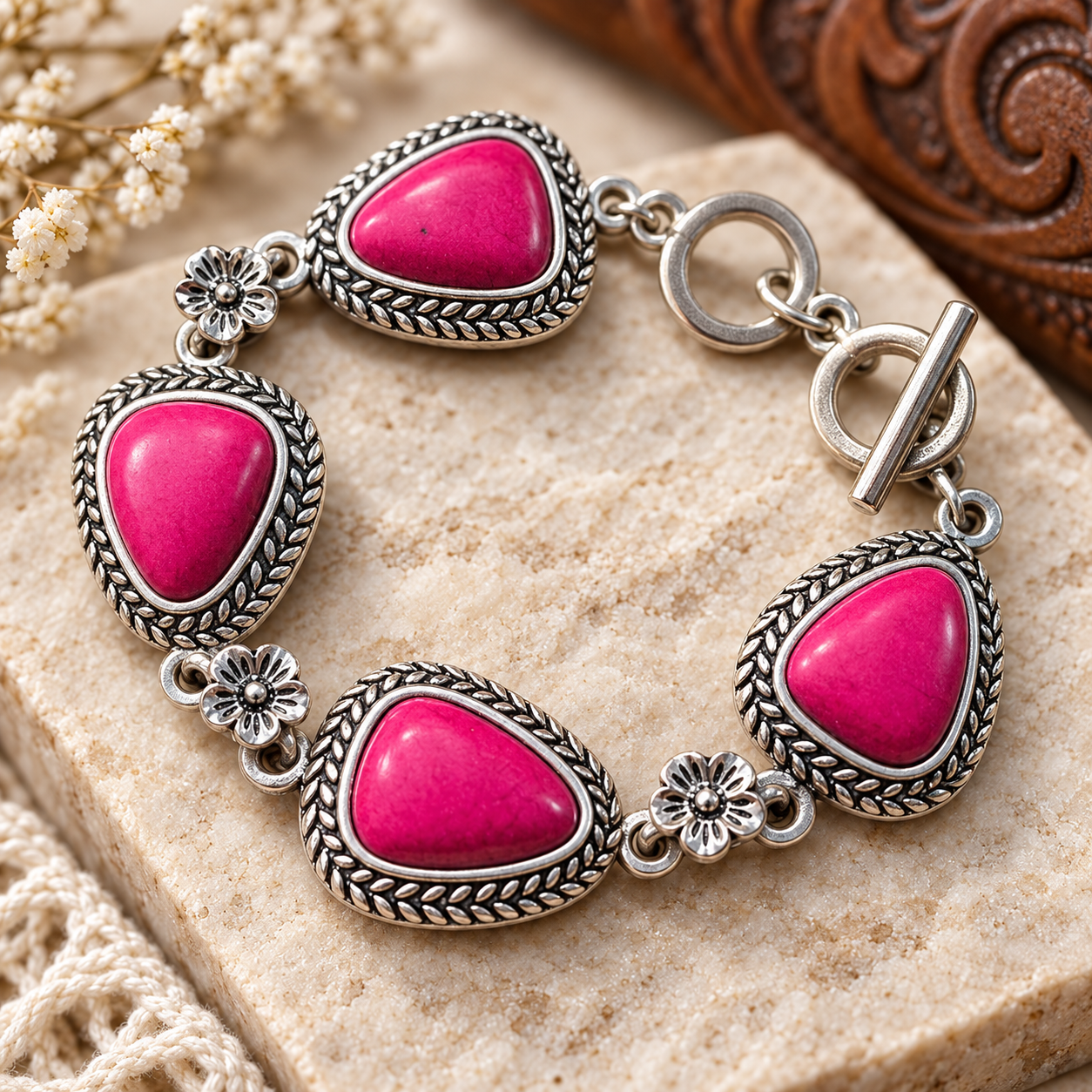 A silver-toned bracelet with five bright pink teardrop-shaped stones, each set in an ornate braided border, displayed on a beige stone surface with dried flowers and a carved wooden object in the background.