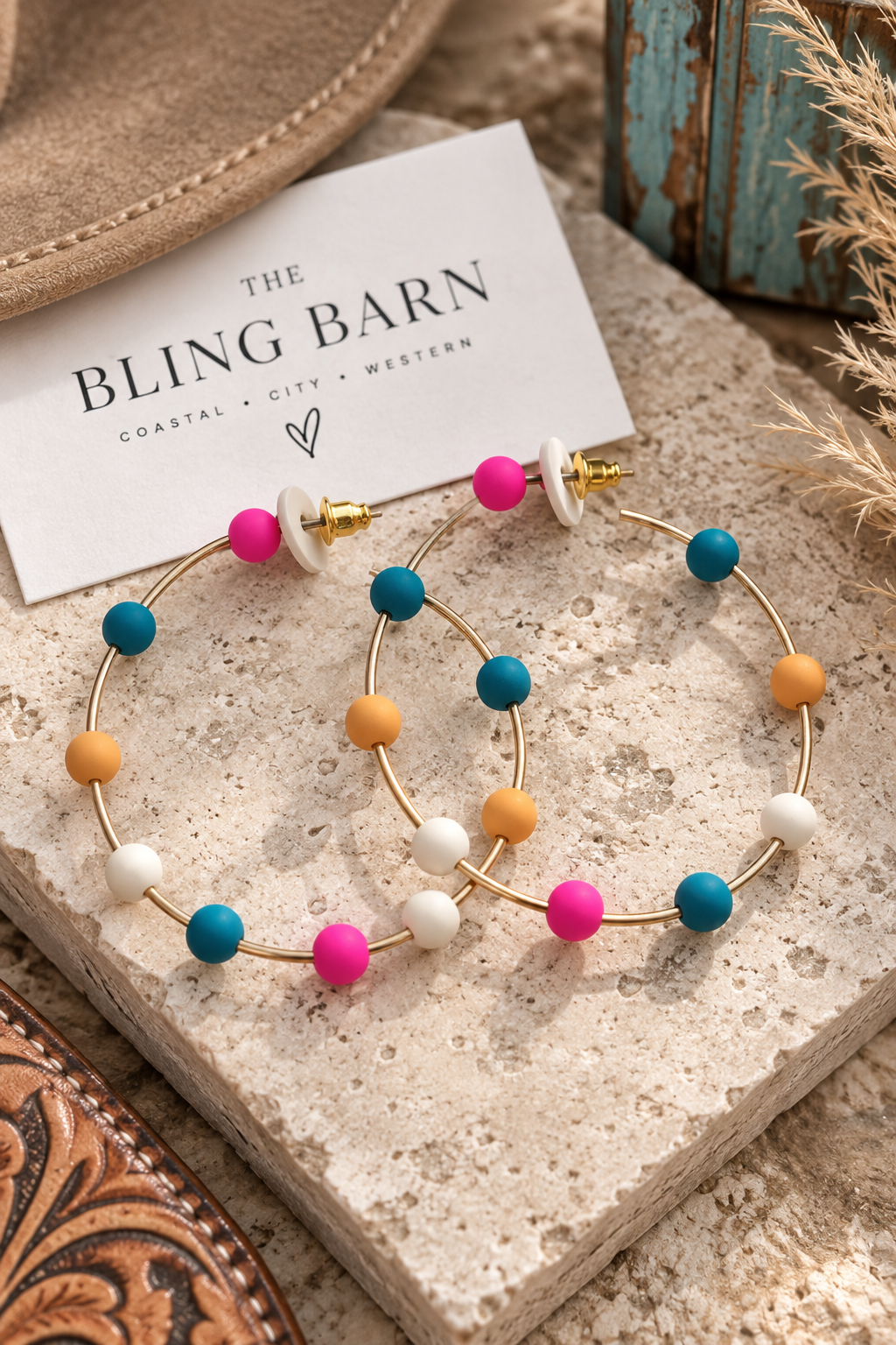 Gold hoop earrings with colorful beaded accents in pink, teal, orange, and white, displayed on a stone slab next to a card reading 'THE BLING BARN'.