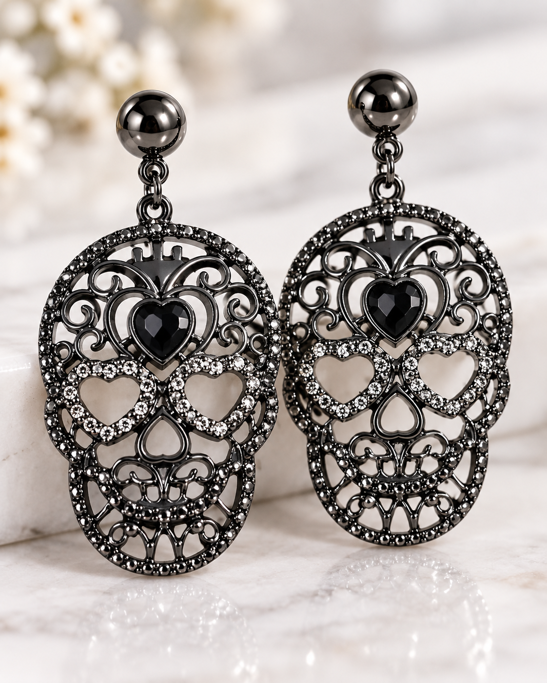 A pair of gothic sugar skull drop earrings with intricate filigree detailing, black heart-shaped crystals, and rhinestone accents, shown on a light marble surface.