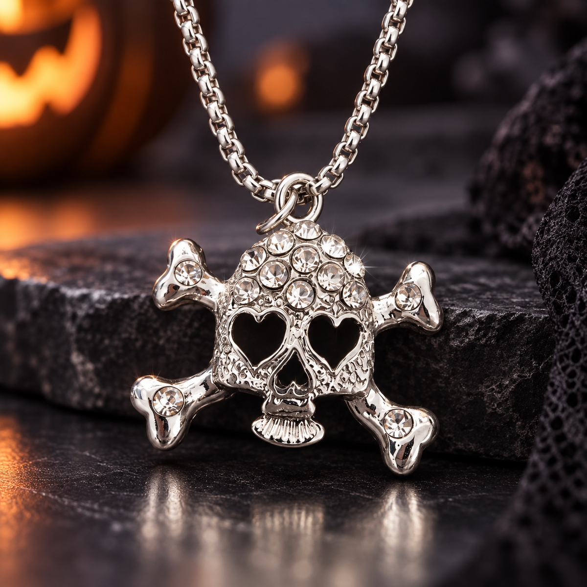 A silver-toned skull and crossbones pendant necklace adorned with clear rhinestones, displayed on a dark textured surface with a blurred Halloween background.