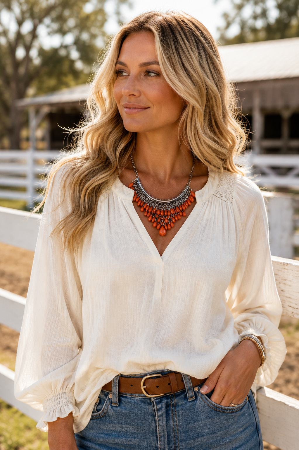 A woman wearing a cream-colored long-sleeve blouse with a V-neckline, paired with blue jeans and a brown belt, standing outdoors in front of a white fence and barn.