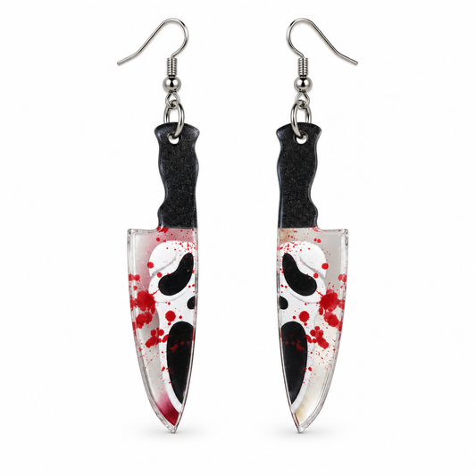 A pair of dangling earrings shaped like knives, featuring a white and black Ghostface mask design with red blood splatter, suspended from silver fishhook wires on a plain white background.