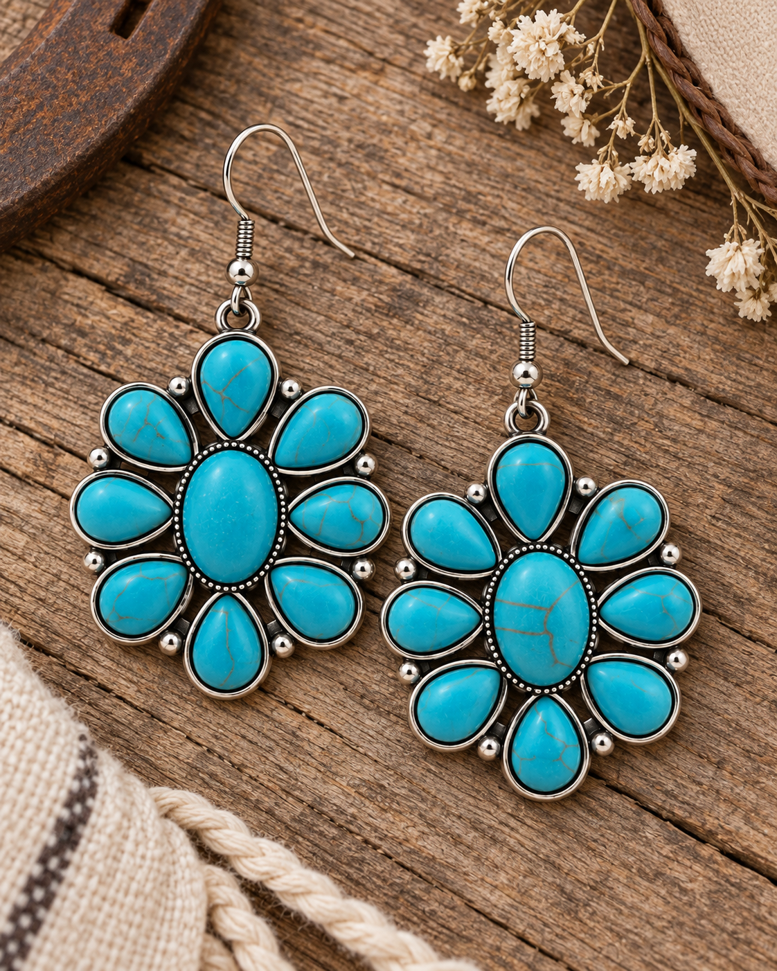 A pair of silver dangle earrings with turquoise stones arranged in a flower-like pattern, displayed on a rustic wooden surface with dried flowers and burlap as props.