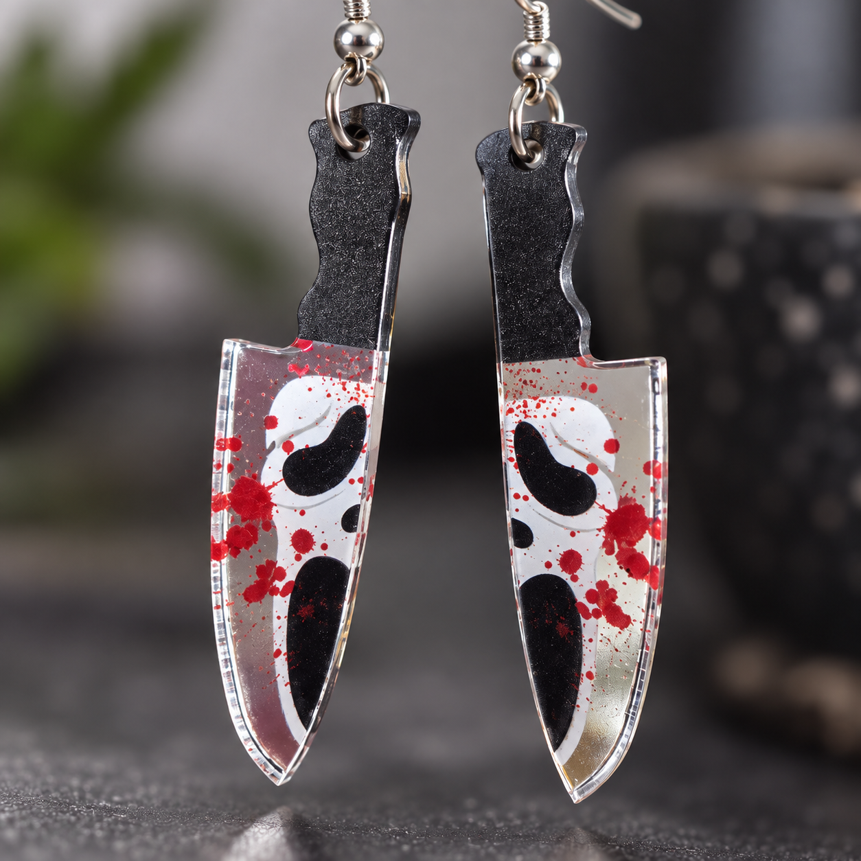 A pair of dangling earrings shaped like kitchen knives, with black handles and white blades decorated with red blood splatter and black eye motifs, shown against a blurred dark background.