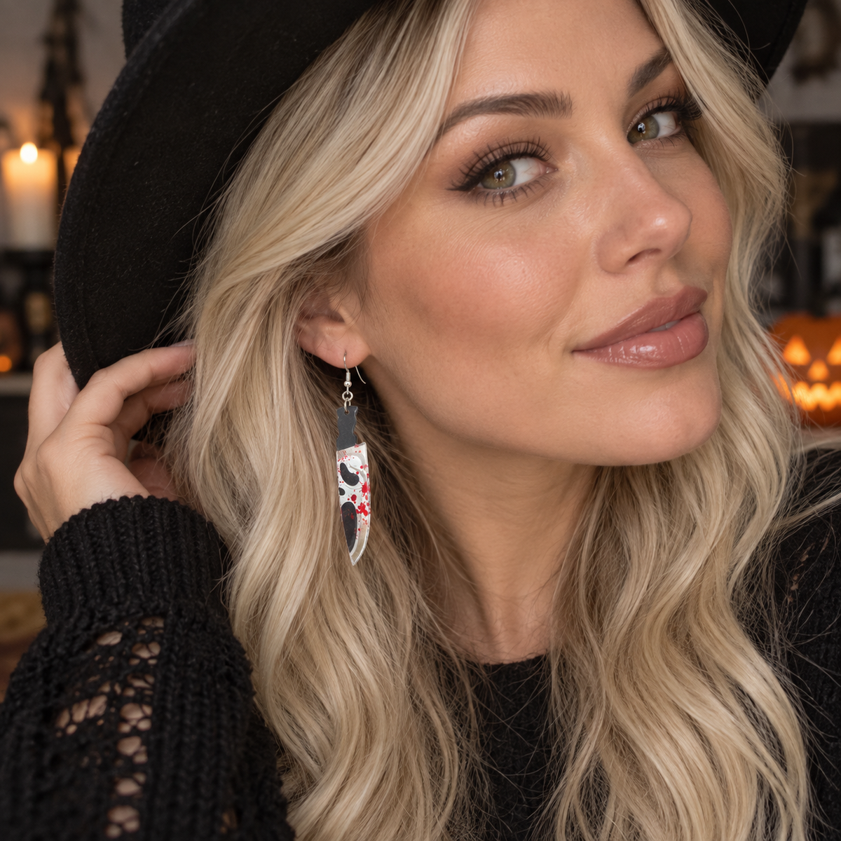 A close-up lifestyle shot of a woman with blonde hair wearing a black hat and black knit sweater, showcasing a long, silver-toned dangle earring decorated with a black cat illustration and red and white star accents.