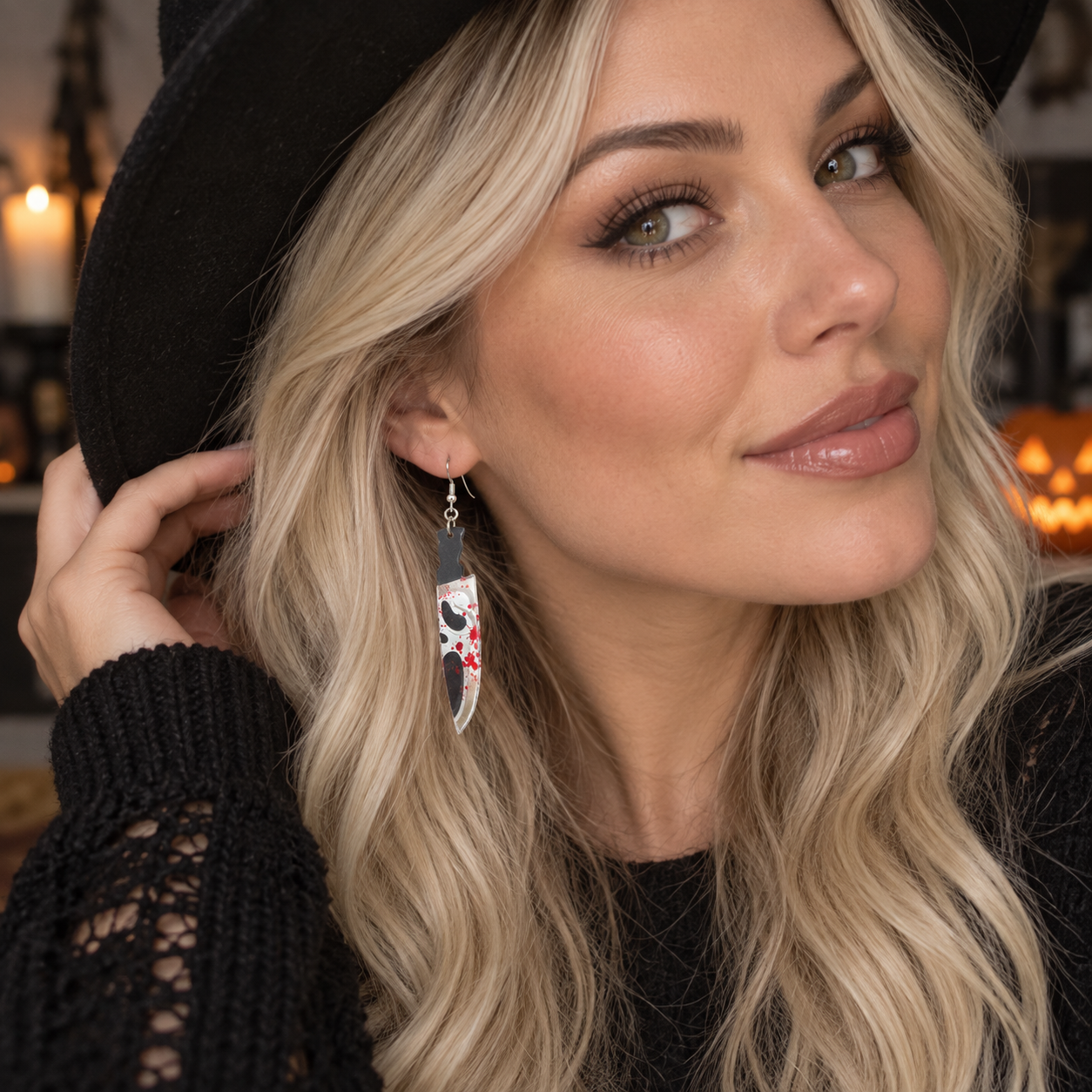 A close-up lifestyle shot of a woman with blonde hair wearing a black hat and black knit sweater, showcasing a long, silver-toned dangle earring decorated with a black cat illustration and red and white star accents.