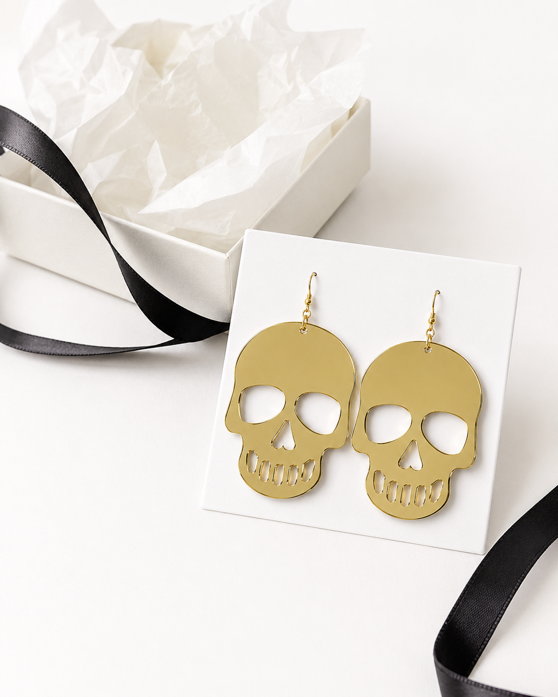 Gilded Afterlife gold skull earrings displayed on a white jewelry card with gift box and black ribbon